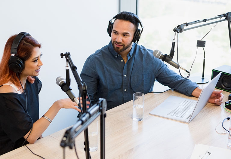 Two podcasters working together in an office with microphones, headphones, and laptop at table
