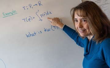 Math professor at white board pointing to equation but facing outward