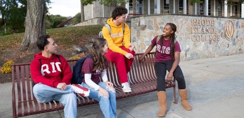Students in RIC gear sitting on a bench, talking, near the RIC sign and library