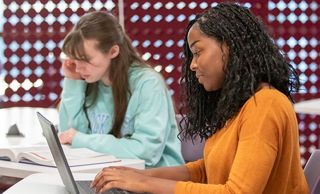 Student typing on laptop, with another student studying nearby